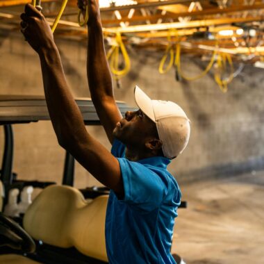 A man wearing a cap works on wiring above a golf cart in a garage.