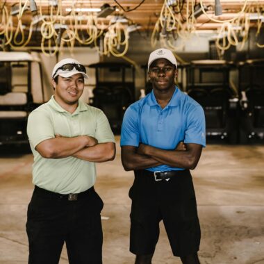 Two men pose confidently in a garage with multiple golf carts. Indoors and energetic.